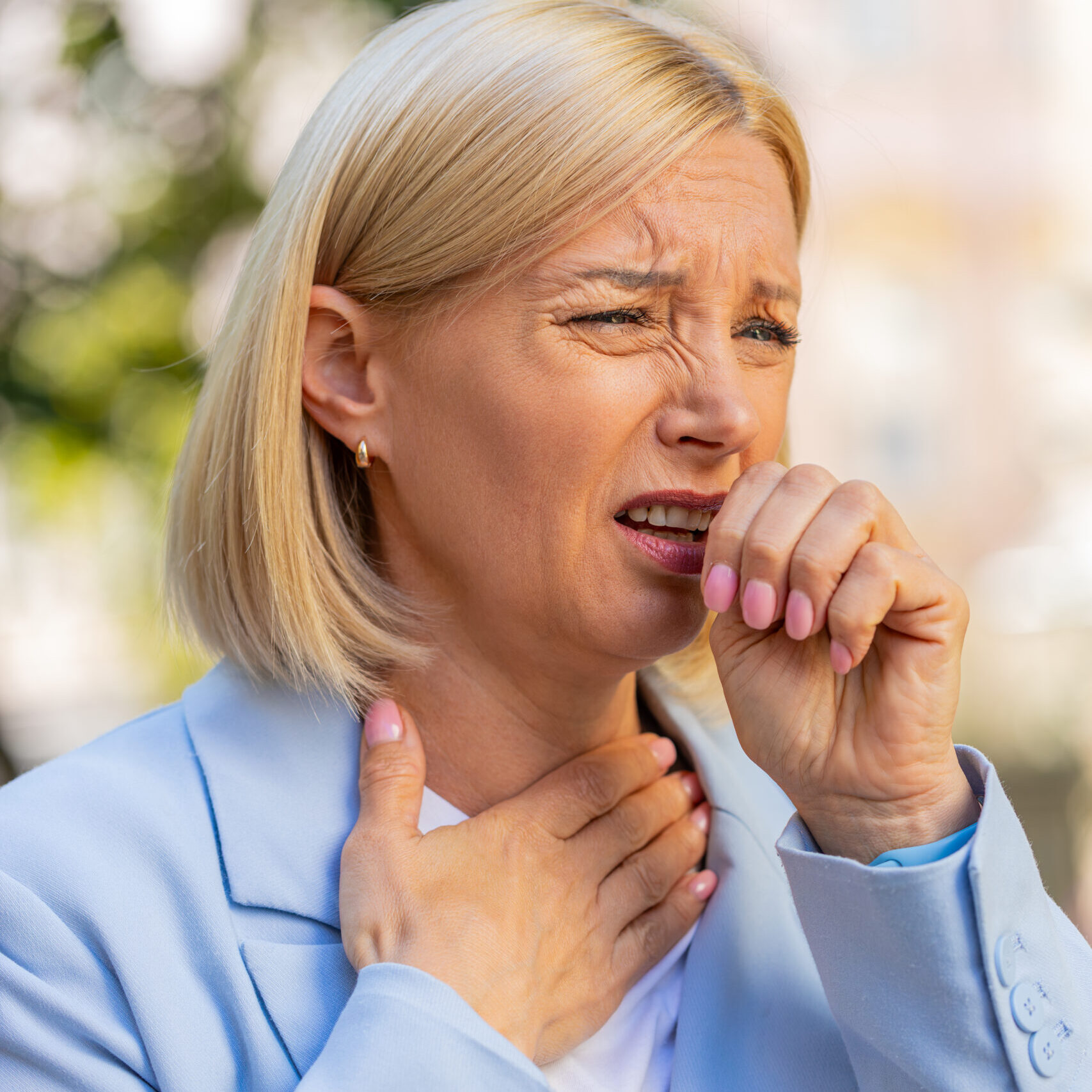 Sick mature businesswoman in formal suit suffering from sore throat while standing on city street. Upset Caucasian female manager with a cold or flu coughing and feels bad. She has a cold or virus.